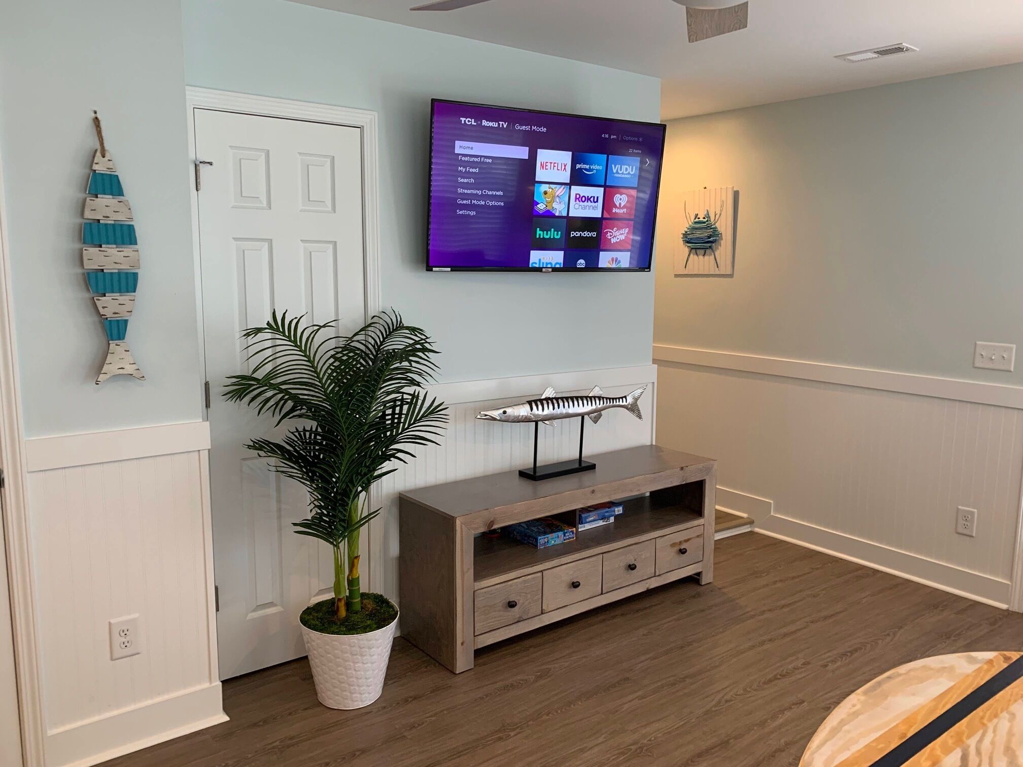 A living room with a flat-screen TV mounted on the wall, a wooden entertainment unit below, a potted plant, and a decorative fish wall art piece.