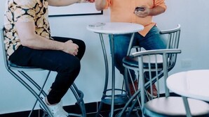 Dos personas sentadas en una mesa redonda en una habitación con un cuadro de un paisaje urbano en la pared, una ventana y una lámpara en el techo.
