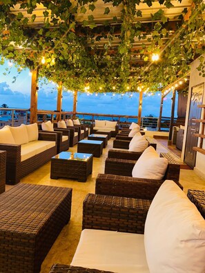 An outdoor seating area with wicker furniture and hanging plants, overlooking a body of water. - Hotel El Reloj (Puerto Plata)