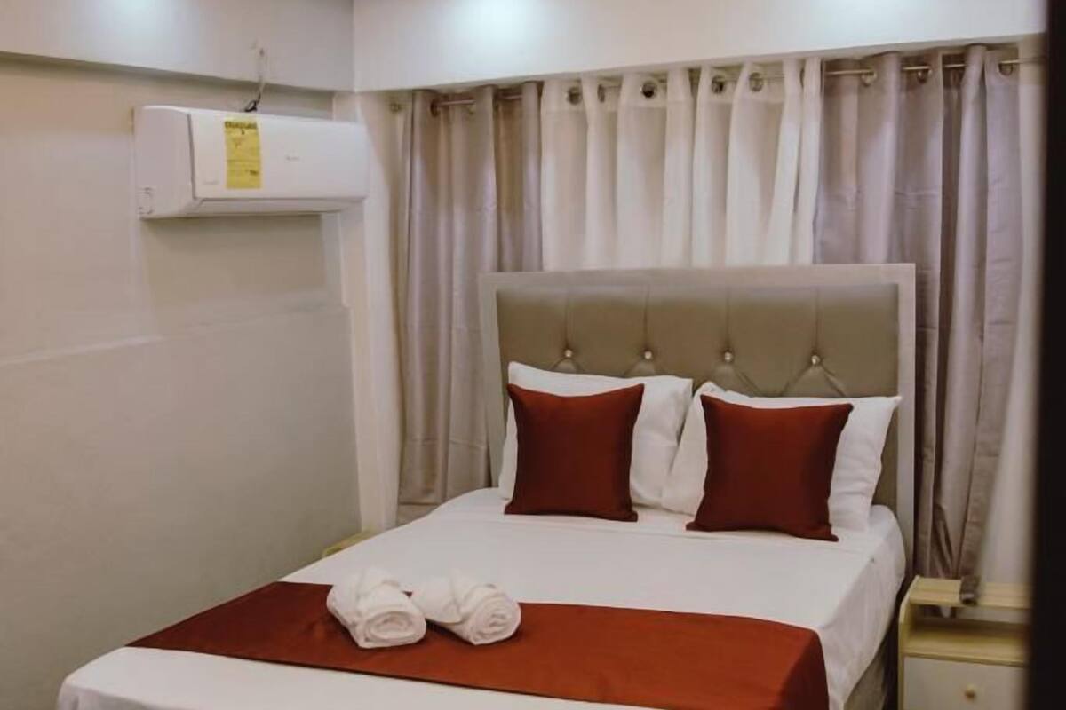 A neatly made bed with a red and white bedspread, a brown upholstered headboard, and a white air conditioning unit mounted on the wall.