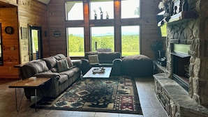 A log cabin interior with a stone fireplace, a leather sofa, a coffee table, and large windows offering a view of the forest. - Gated Hilltop Luxury Lodge on 100 acres - Relax and Enjoy AMAZING VIEWS (Smithville)