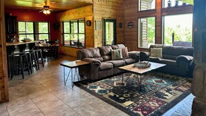 A log cabin interior with a dining area, a living room with a sofa and coffee table, and a large window with a view of the outdoors. - Gated Hilltop Luxury Lodge on 100 acres - Relax and Enjoy AMAZING VIEWS (Smithville)