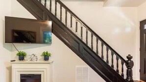 A staircase with a black handrail and balusters, a white wall, a fireplace with a TV above it, and a door to the right. - Northfork Townhouse walk to all (Greenport)