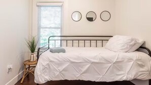 A bedroom with a bed, a small side table with a plant, and a window with blinds. - Northfork Townhouse walk to all (Greenport)