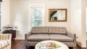 A living room with a grey sofa, a round coffee table with magazines, a wooden side table, and a large mirror on the wall. - Northfork Townhouse walk to all (Greenport)