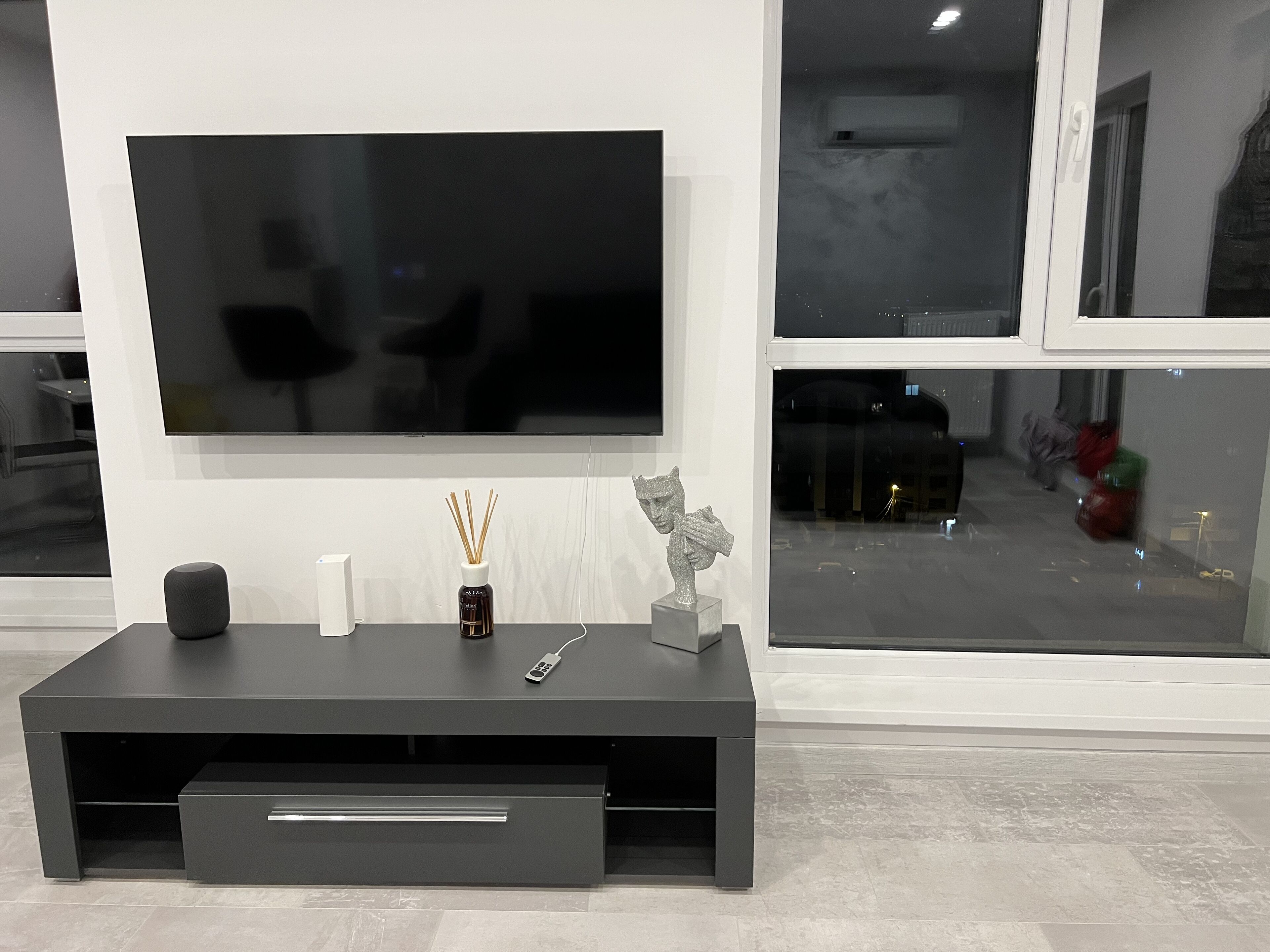 A modern living room with a large flat-screen TV mounted on a white wall, a dark grey entertainment unit with a drawer, a small vase with reed sticks, and a sculpture.