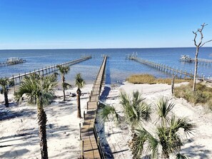 Une plage avec une promenade en bois qui mène à l’eau, entourée de palmiers et surplombée d’un ciel bleu clair.