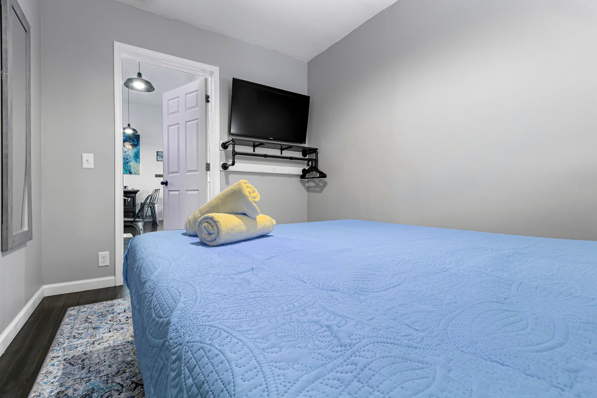 A bedroom with a blue quilted bedspread, a rolled-up yellow towel, a mounted TV, and a door leading to another room.