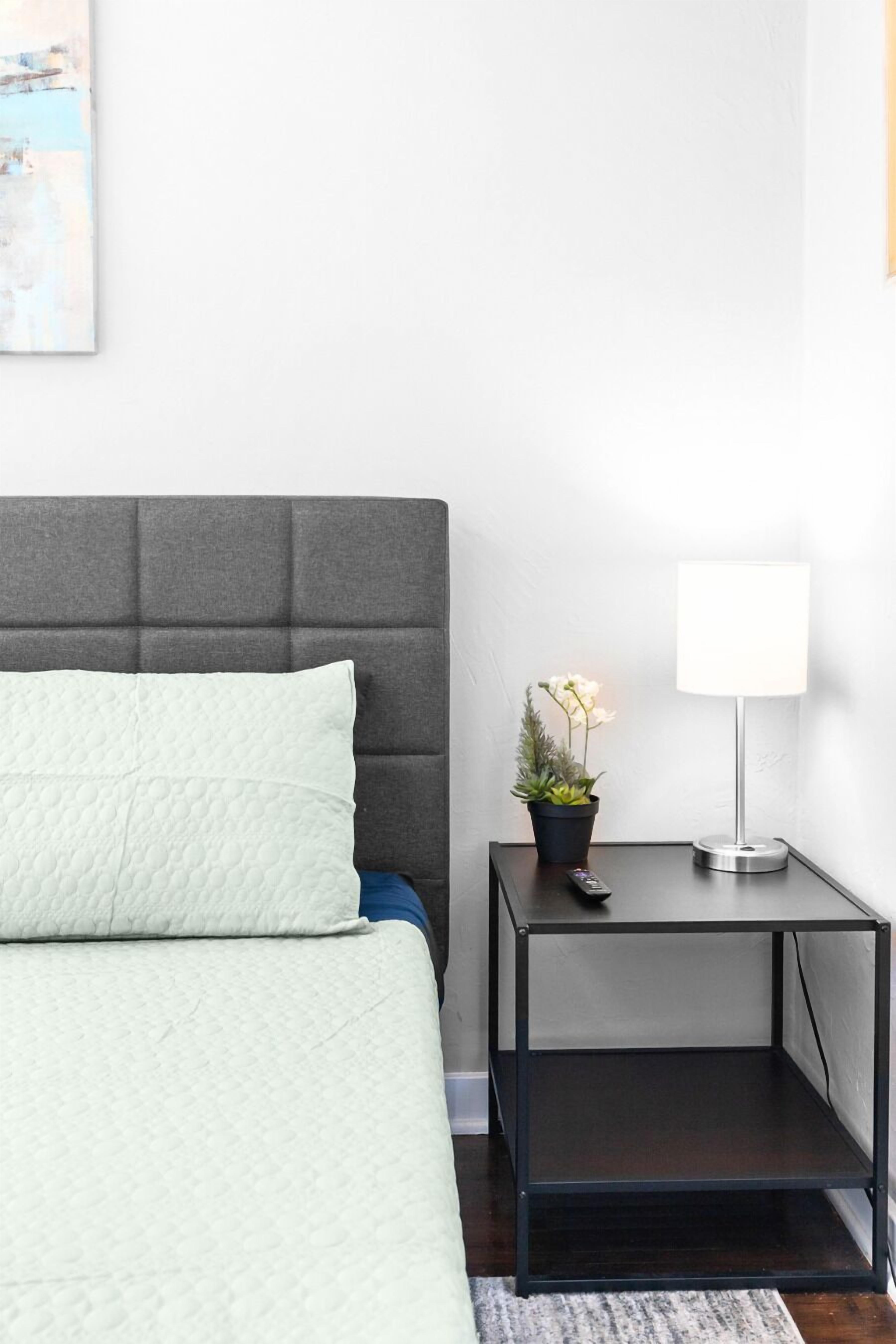 A neatly made bed with a grey headboard and a single white pillow. A small black nightstand with a white lamp and a potted plant is beside the bed.