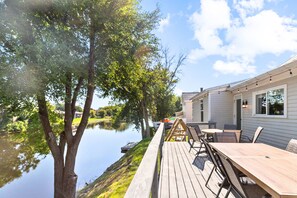 Terraza de madera con mobiliario exterior con vistas a un lago tranquilo, conectada a un edificio con amplios ventanales.