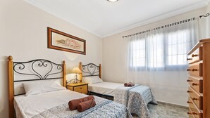 Ein Hotelzimmer mit zwei Betten, einem Nachttisch mit Lampe, einer Holzkommode und einem Fenster mit durchsichtigen Vorhängen.