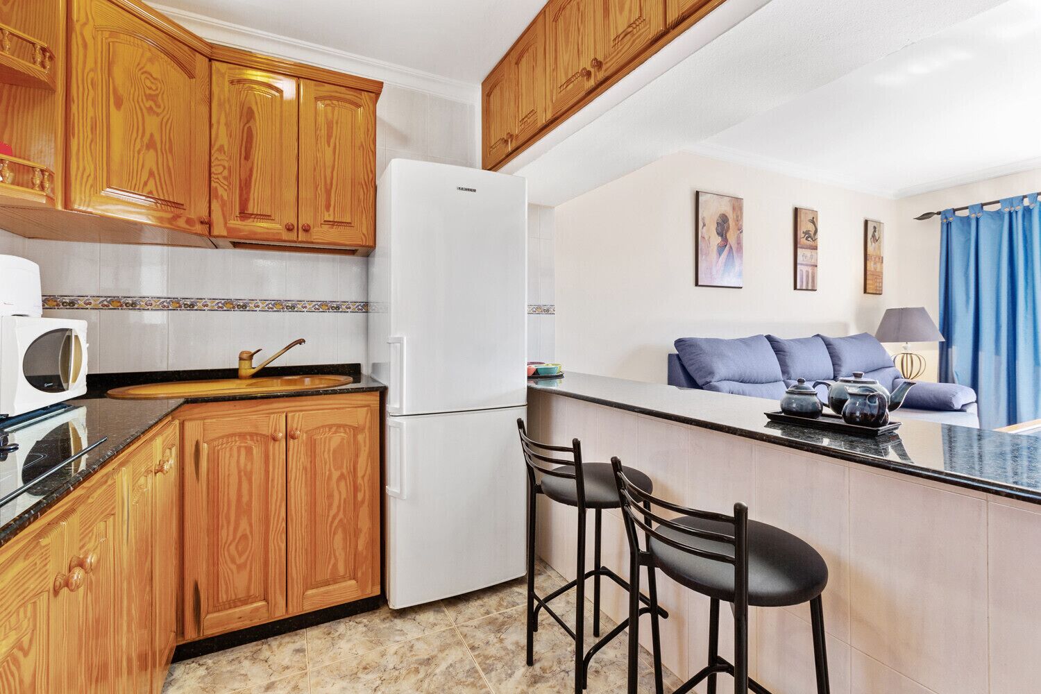 A compact kitchen with wooden cabinets, a white refrigerator, a microwave, and a small bar area with stools.