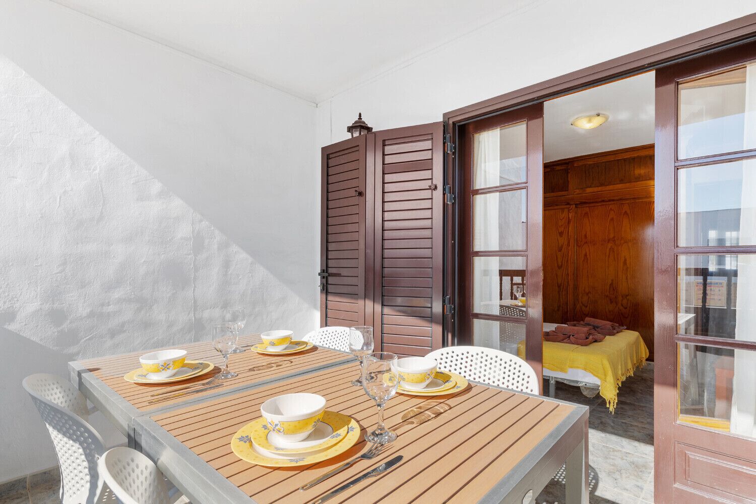 Un comedor con una mesa de madera dispuesta para cuatro, sillas blancas y vistas a una habitación con una cama y paneles de madera.