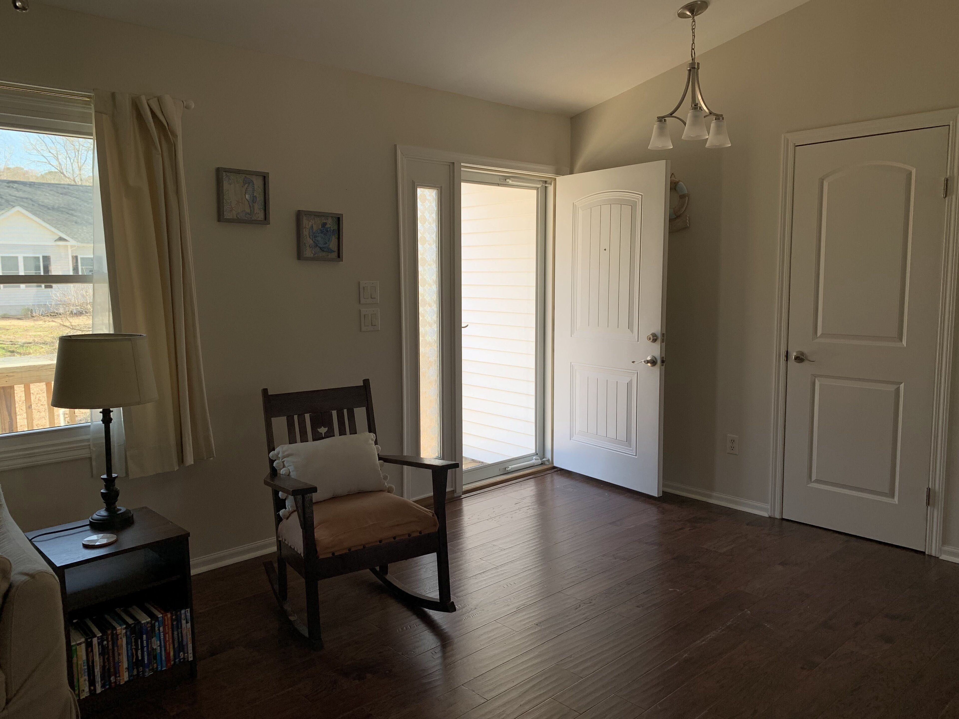 Una habitación con piso de madera, un sillón mecedor, una estantería, una lámpara y una puerta que da a un espacio exterior.