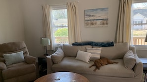 Una sala de estar con un sofá color beige, un sillón marrón, una mesa de centro de madera y un perro acostado en el sofá.