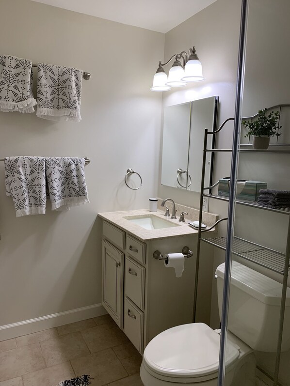 A bathroom with a toilet, sink, and mirror. There are towels hanging on the wall and a shelf with toiletries. - Open-spaced Beach House - 3 Bed, 2 Bath (Ocean View)