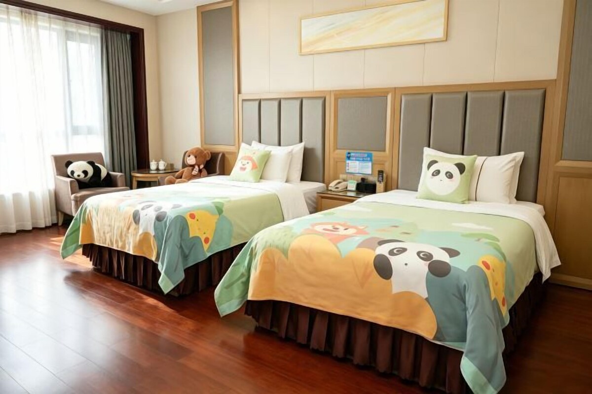 A hotel room with two beds, each with a panda-themed bedspread, a wooden floor, a chair with a panda cushion, and a window with curtains.