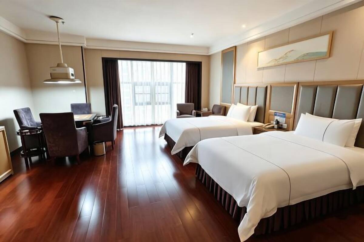 A hotel room with two beds, a desk, a chair, a lamp, and a large window.