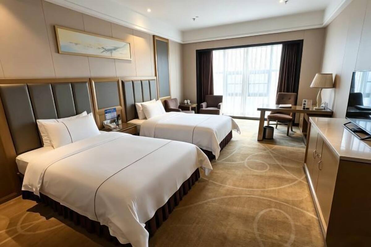 A hotel room with two beds, a desk, a chair, a TV, and a large window.