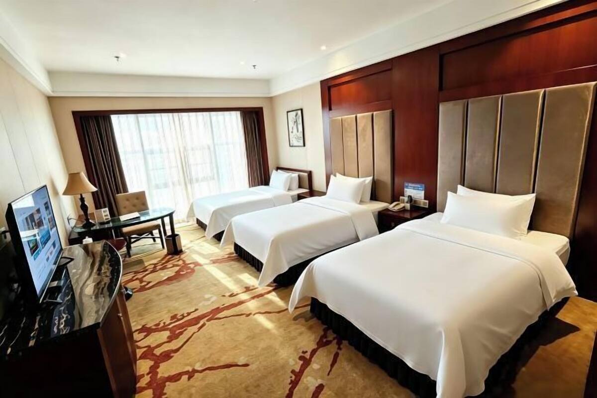 A hotel room with three beds, a desk with a computer, and a large window with curtains.