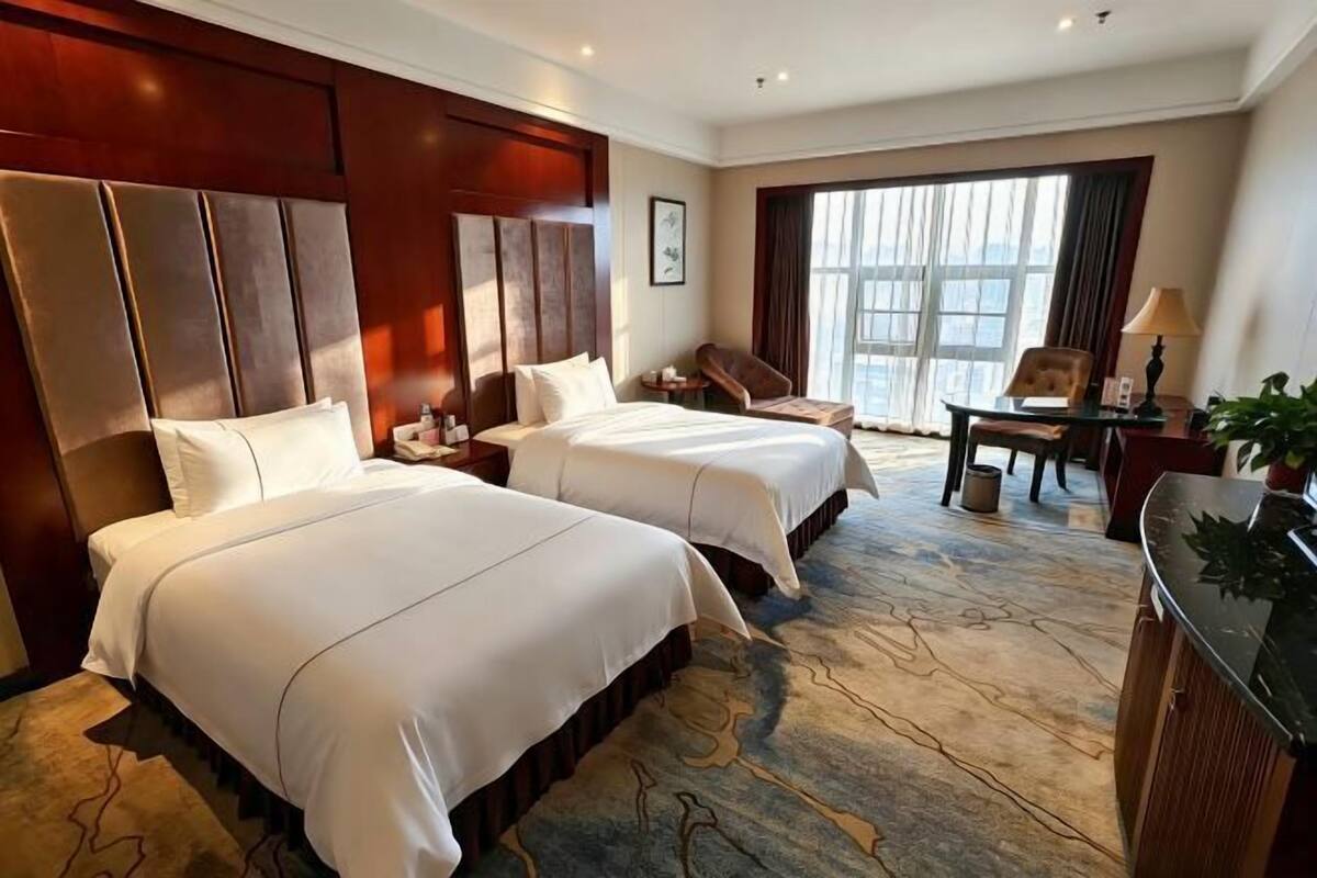 A hotel room with two beds, a desk, a chair, and a large window.