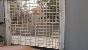 A gated entrance with a metal fence, a concrete planter with a plant, and a paved area leading to a building with a tiled roof.