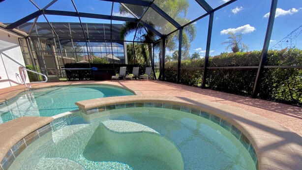 A swimming pool with clear water, surrounded by a covered patio area with chairs and a view of palm trees.