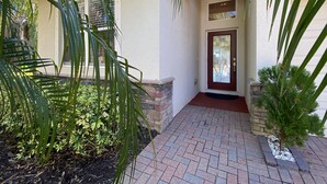 A house entrance with a red door, a brick pathway, and surrounding greenery.