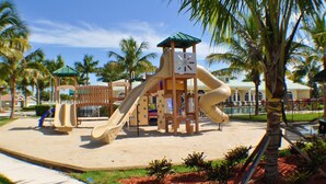 A playground with slides and climbing structures, surrounded by palm trees and a building in the background.