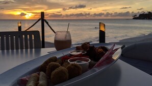 Une table avec une assiette de nourriture et une boisson, donnant sur un coucher de soleil sur l’océan.