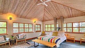 Une chambre confortable avec un grand lit, un banc et une chaise. Il y a des fenêtres donnant sur des arbres et un ventilateur de plafond.