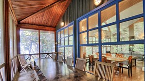 Une salle à manger spacieuse avec une longue table et des chaises en bois, de grandes fenêtres aux cadres bleus et une vue sur la forêt environnante.