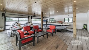 A covered deck with wooden flooring, red and black seating, and a boat docked outside. - SW Exposure at this 4 bdrm, 3 bath cottage with stunning views of Lake Muskoka! (Gravenhurst)