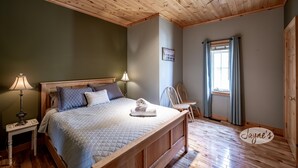 A bedroom with a wooden bed, a nightstand with a lamp, a window with curtains, and a wooden door. - Unique 8 bed 5bath cottage on the Joe River! (Port Carling)