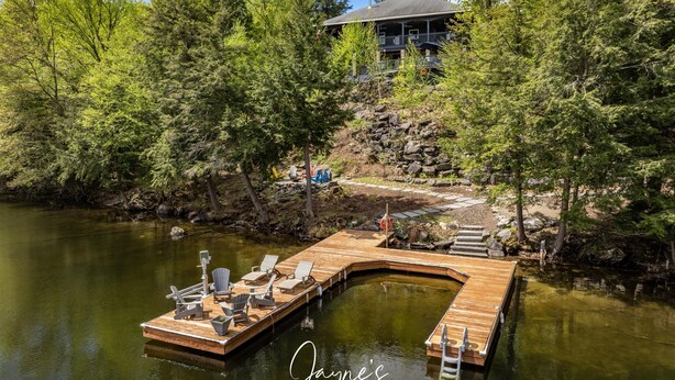 A wooden dock with chairs and a small boat, surrounded by trees and a house in the background. - Unique 8 bed 5bath cottage on the Joe River! (Port Carling)