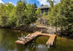 A wooden dock with chairs and a small boat, surrounded by trees and a house in the background.