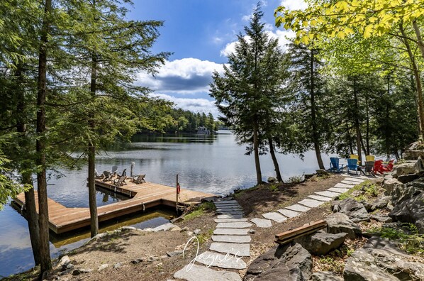 A wooden dock extending into a calm lake surrounded by trees, with a stone path leading to a docked boat.