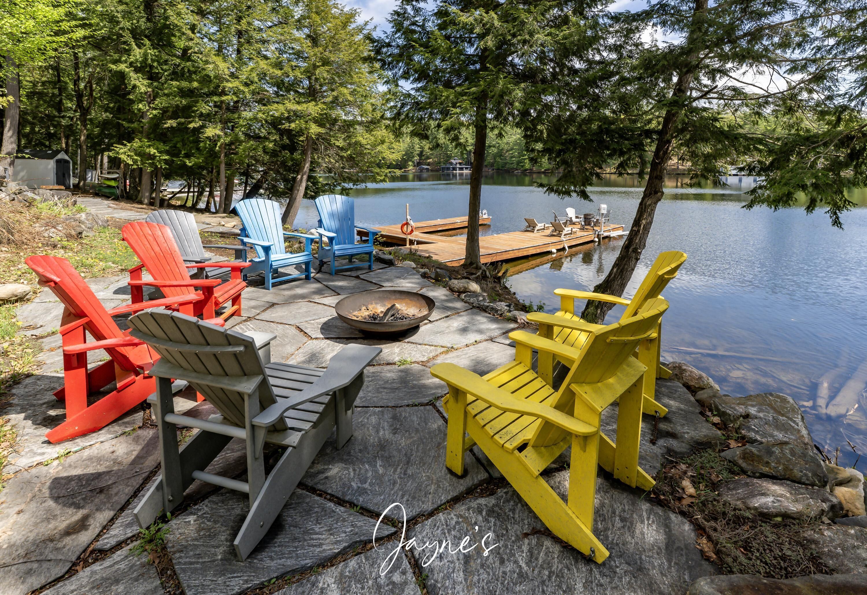 A lakeside seating area with Adirondack chairs in red, blue, gray, and yellow, a fire pit, and a dock extending into the water.