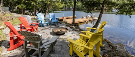 A lakeside seating area with Adirondack chairs in red, blue, gray, and yellow, a fire pit, and a dock extending into the water.