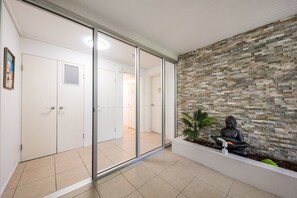 A hallway with a glass door leading to a room, a stone wall with a Buddha statue, and a tiled floor. - Ocean Dreams Studio Apartment On Airlie (Airlie Beach)