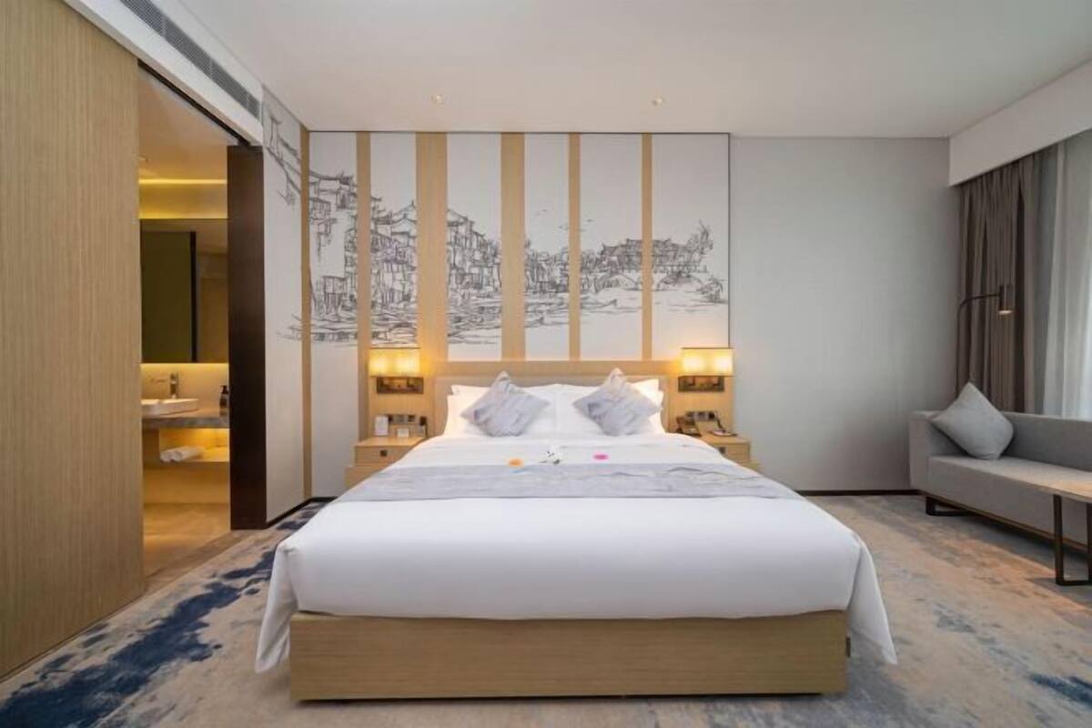 A modern hotel room with a large bed, bedside tables, a sofa, and a wall with a large artwork.