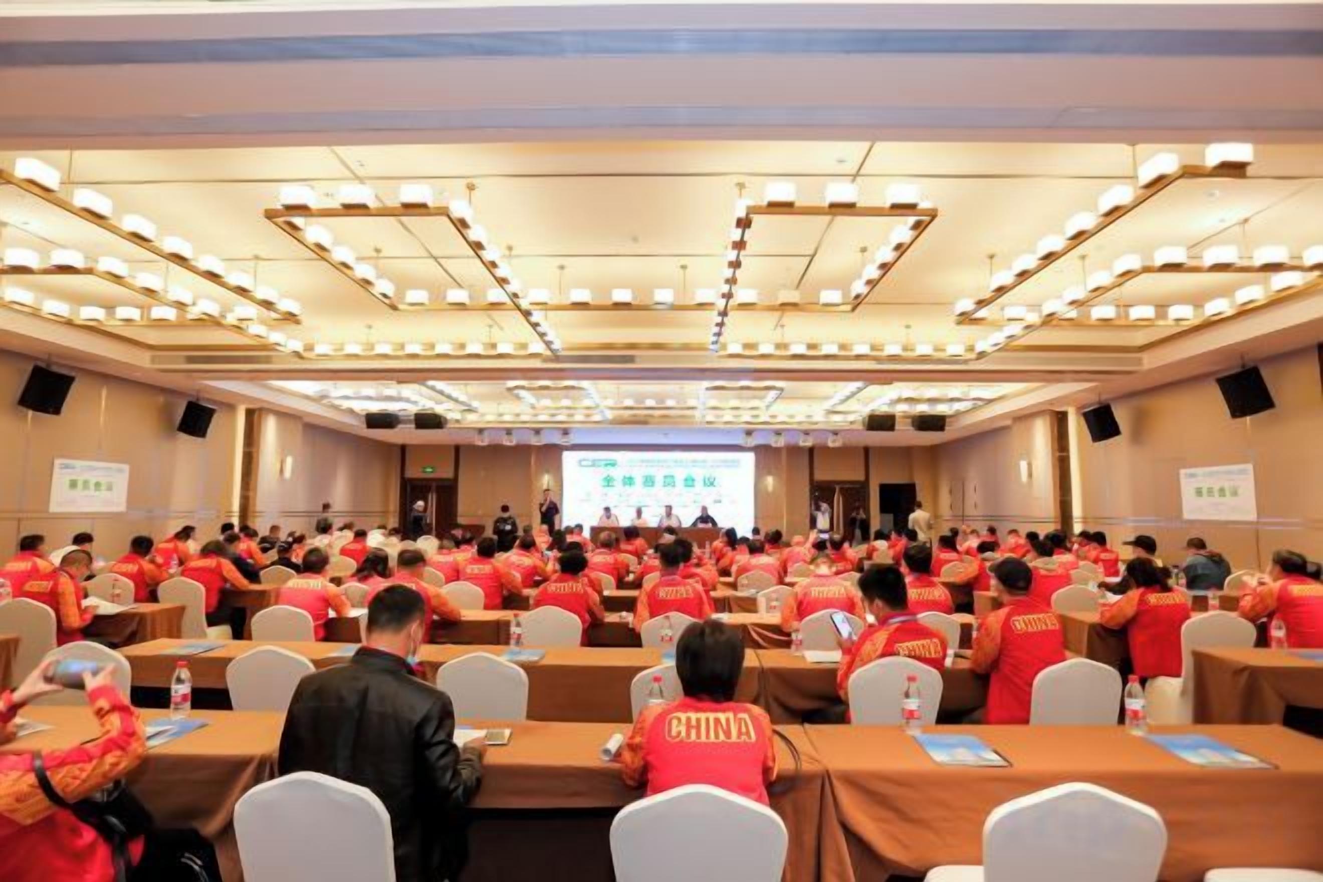 Uma sala de conferências com fileiras de mesas e cadeiras, uma tela de apresentação na frente e participantes uniformizados de vermelho.