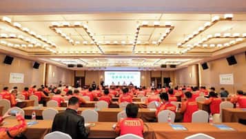 Uma sala de conferências com fileiras de mesas e cadeiras, uma tela de apresentação na frente e participantes uniformizados de vermelho.