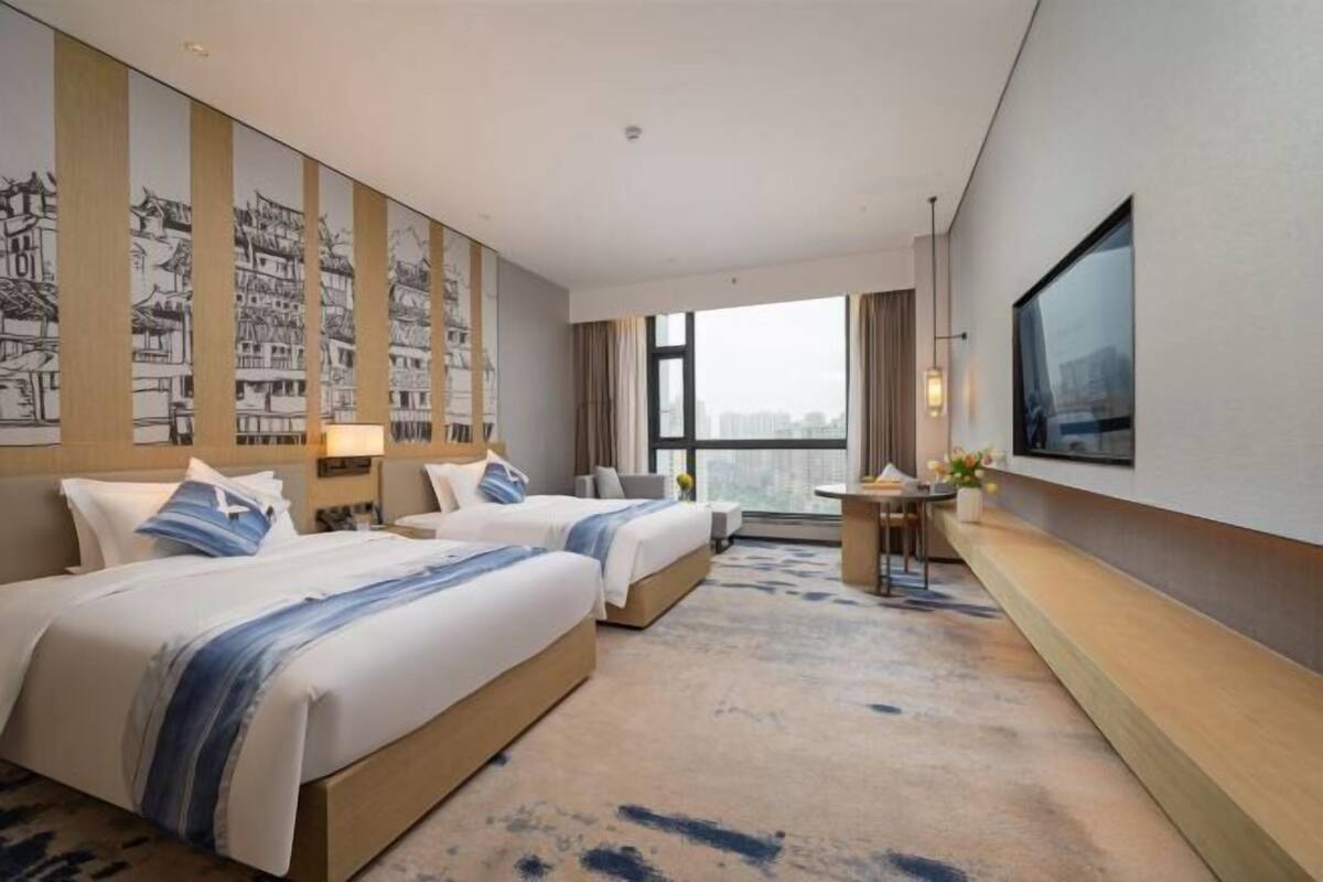 A hotel room with two beds, a desk, a TV, and a large window.
