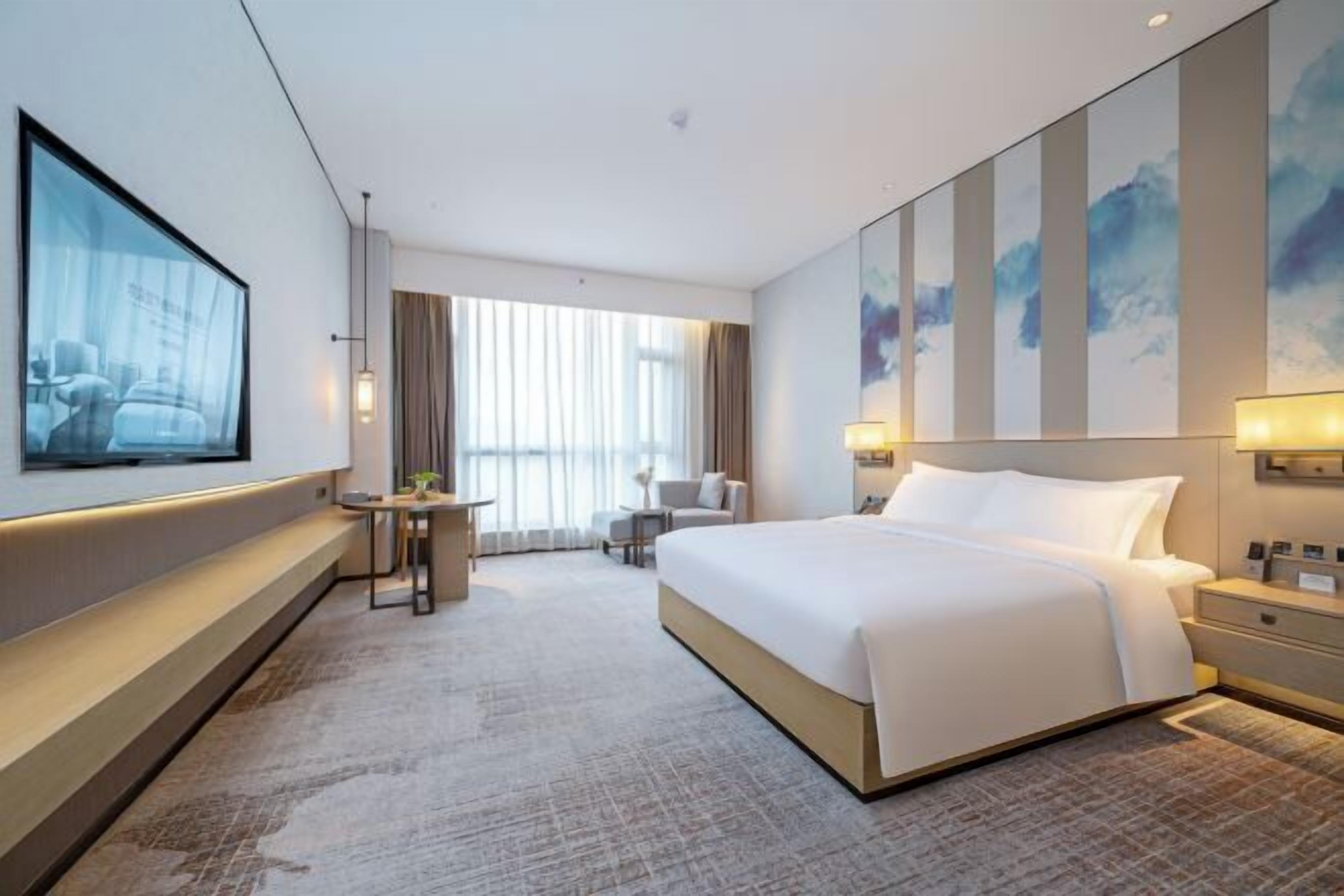 A hotel room with a large bed, a TV mounted on the wall, a desk with a chair, and a window with curtains.