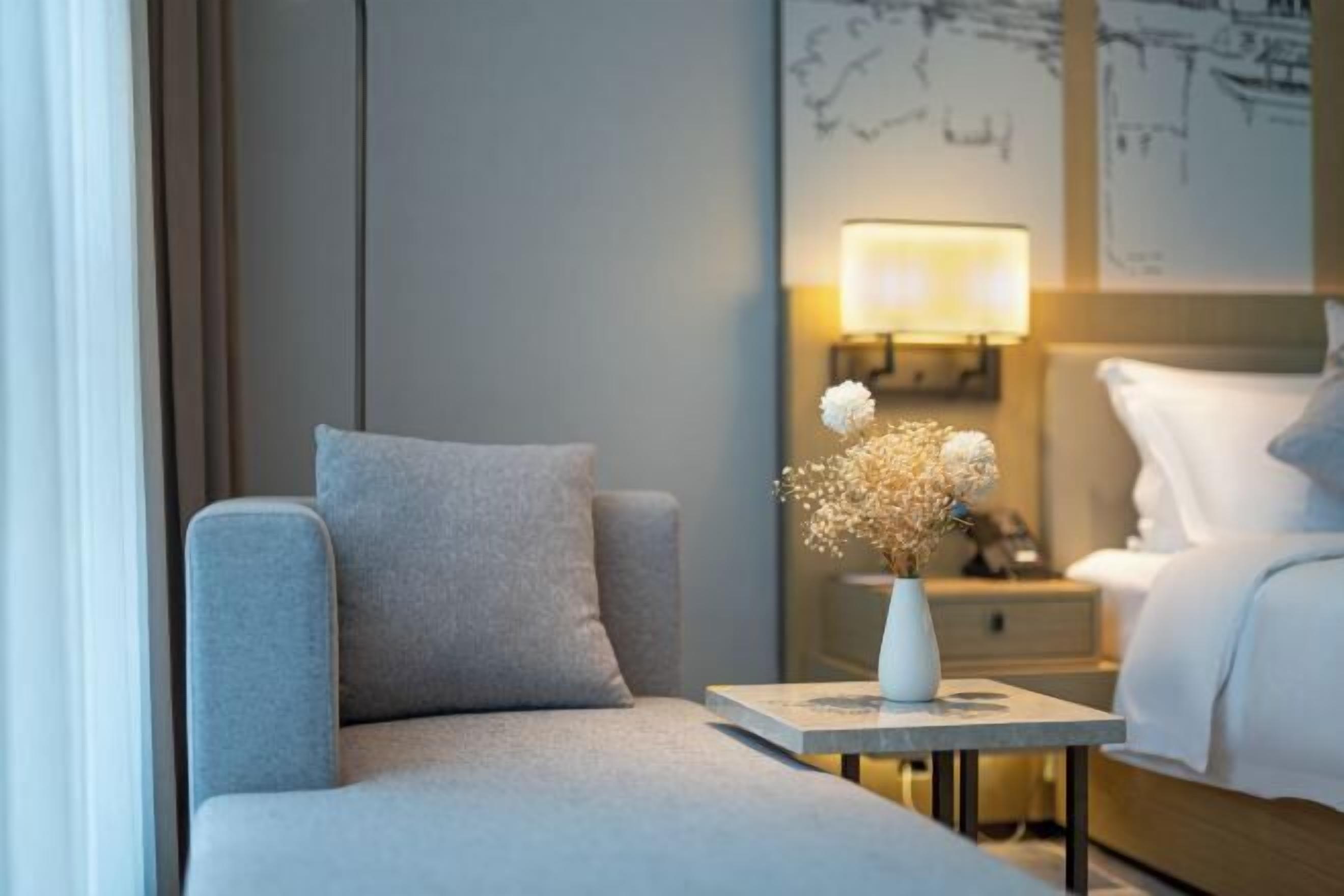 A hotel room with a sofa, a bedside table with a vase of flowers, a lamp, and a bed with white bedding.