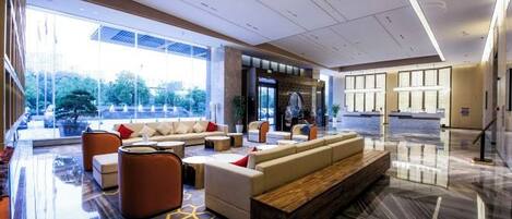 A modern hotel lobby with a seating area, a reception desk, and large windows offering a view of the outdoors.