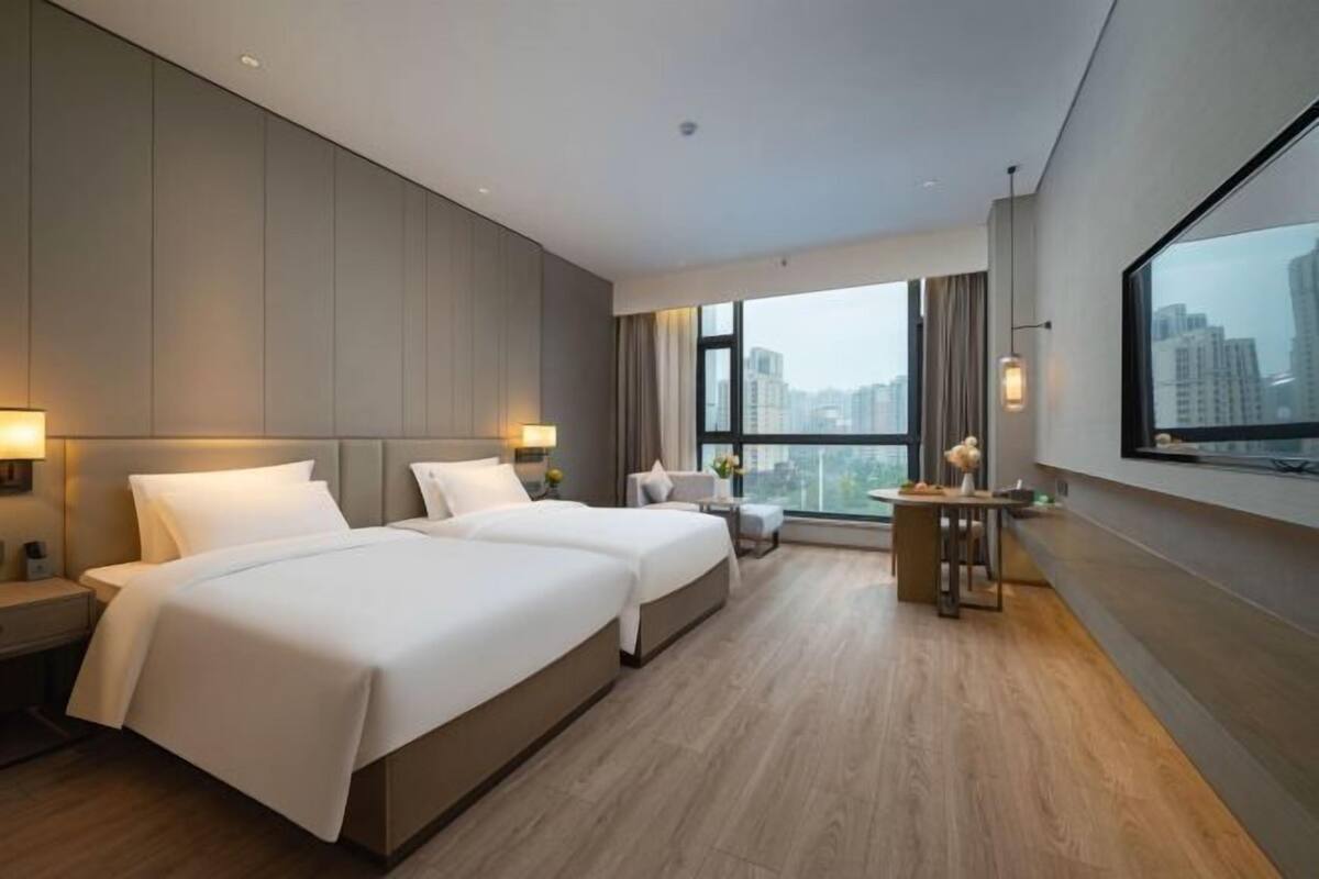 A hotel room with two beds, a small table, a chair, and a large window offering a city view.