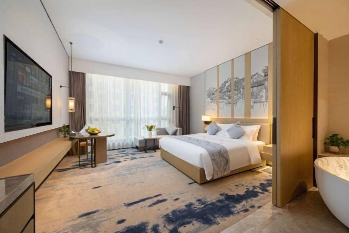 A modern hotel room with a large bed, a desk, a chair, a small table with a plant, and a large window with sheer curtains.