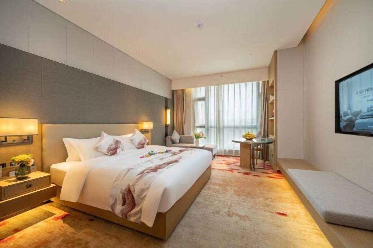 A hotel room with a large bed, bedside tables, a TV, a small table with chairs, and a window with curtains.